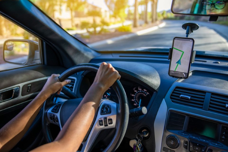 A woman driving a car with a smartphone in a mobile holder displaying a car GPS tracker app, monitoring the location of another vehicle in real-time.